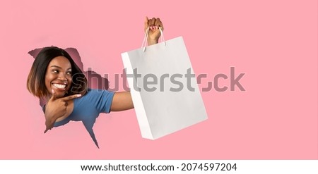 Shopping And Sales. Excited young black woman holding paper shopper bag in hand with mock up for your brand logo design, breaking through hole in slit pink paper, pointing at empty space for advert