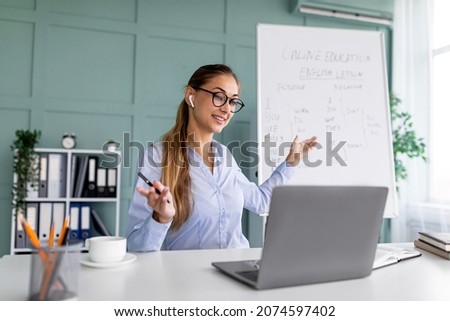 Online education. Young English teacher having video conference chat with students, talking to laptop webcamera. Woman wearing glasses and wireless earphones, explaining grammar rules Royalty-Free Stock Photo #2074597402