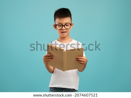 Portrait Of Little Asian Schoolboy In Eyeglasses Reading Book While Standing Isolated Over Blue Studio Background, Nerdy Preteen Korean Male Kid Enjoying Study And Learning Process, Copy Space