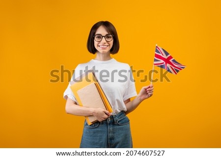 Travel and study, learn English language. Zoomer smiling woman with notebooks and small flag go to university, college or school, isolated on orange background, studio shot Royalty-Free Stock Photo #2074607527