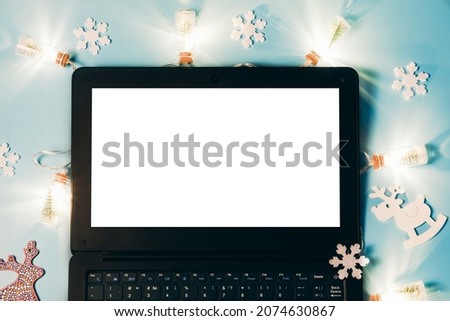 Christmas flatlay with a garland of bottles, a toy wooden deer and white snowflakes on a blue background. New Year's laptop with a white monitor, copy space, mock up.