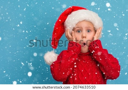 Funny surprised child in Santa hat and red sweater on blue snowy background. 