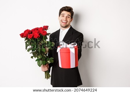 Concept of holidays, relationship and celebration. Image of handsome smiling guy in black suit, holding bouquet of red roses and giving you a gift, standing against white background