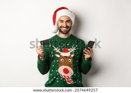 New Year, online shopping and christmas concept. Happy bearded man buying in internet with credit card and phone, wearing Santa hat and sweater, white background