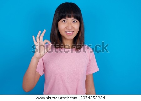 Glad attractive young asian woman wearing pink t-shirt against blue background shows ok sign with hand as expresses approval, has cheerful expression, being optimistic.
