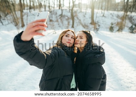 Two girlfriends in warm clothes take a selfie on a smartphone while walking in a winter park, looking at the camera and posing.