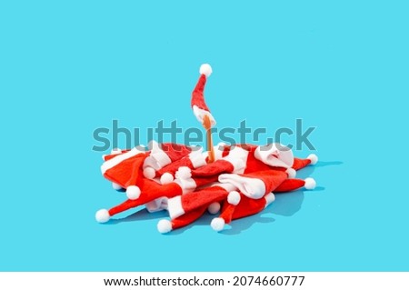 A pile of red Santa Clause's hats with white pompoms and an arm emerging with one red hat. Pastel blue background. Surreal Christmas or New Year's festivities creative concept. Banner or advertisement