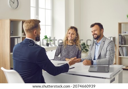 Manager meeting clients in his office. Happy young married couple sitting at table listening to realtor, real estate agent, loan broker or mortgage advisor telling about options they can choose from Royalty-Free Stock Photo #2074660939
