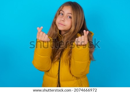 brunette kid girl wearing yellow jacket over blue background doing money gesture with hands, asking for salary payment, millionaire business