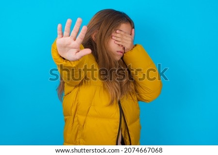 brunette kid girl wearing yellow jacket over blue background covers eyes with palm and doing stop gesture, tries to hide from everybody.