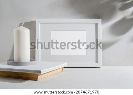 Empty white frame with a passe-partout, a white candle on a concrete plate, and a stack of books in a light interior. Mockup with a place for your design, illuminated by sunlight.