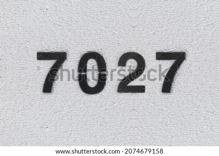 Black Number 7027 on the white wall. Spray paint. Number seven thousand twenty seven.