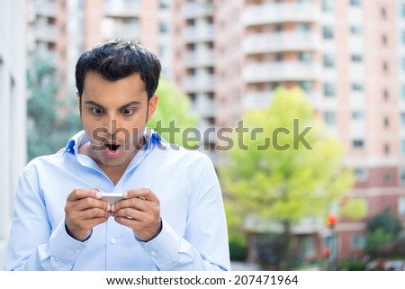 Closeup portrait, funny young man, shocked surprised, wide open mouth, by what he sees on his cell phone, isolated outdoors building background. 