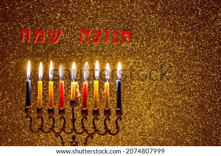 Gold menorah and burning wax candles, festive blurred background with Hebrew letters translated as Happy Hanukkah