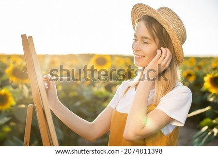 Portrait of a woman who is standing in a field of sunflowers and looking at the picture. The canvas is standing on an easel. She is smiling and holding a brush. She is adjusting her hair.