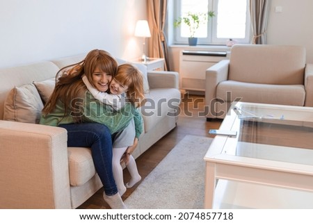 Beautiful woman and her charming little daughter are hugging and smiling. Smiling single young mum embracing little daughter with toy, playing in living room at home, mother laughing with child
