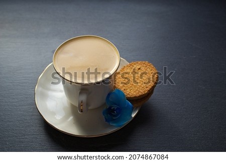 A cup of coffee over black background. Blue orchid, white coffee, biscuit, digestive. Picture with space for text.