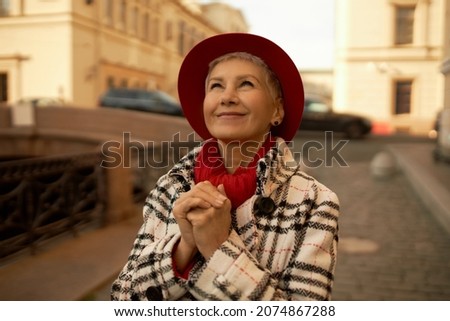 Picture of charming 50-years-old extraordinary female in red stylish hat dressed in plaid coat standing on street with grateful face expression, thanking God or Universe for opportunity to travel