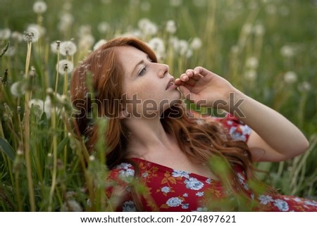 Red-haired girl in a red dress in a field among fading dandelions