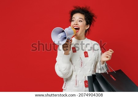 Young woman in white sweater with tags sale in store showroom hold package bags with purchases after shopping scream in megaphone announces discounts Hurry up isolated on plain red background studio