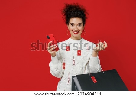 Young cheerful happy female costumer woman wear white knitted sweater with tags sale hold package bags with purchases after shopping using mobile cell phone isolated on plain red background studio.