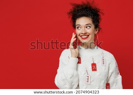 Young female costumer woman 20s wear white knitted sweater with tags sale in store showroom talk speak on mobile cell phone conducting pleasant conversation isolated on plain red background studio.