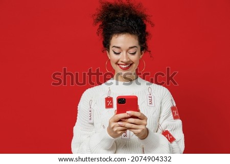 Young smiling happy caucasian female costumer woman 20s wear white knitted sweater with tags sale in store showroom hold in hand use mobile cell phone isolated on plain red background studio portrait
