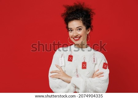 Young smiling cheerful satisfied happy female costumer woman 20s wear white knitted sweater with tags sale in store showroom hold hands crossed folded isolated on plain red background studio portrait