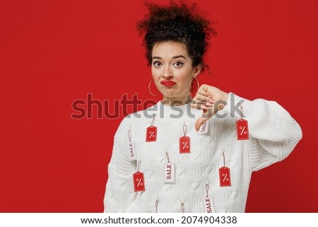 Young sad displeased unhappy female costumer woman 20s wearing white knitted sweater with tags sale in store showroom show thumb down dislike gesture isolated on plain red background studio portrait
