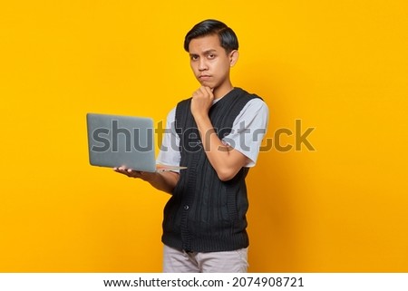 Portrait of confident Asian man holding laptop and looking at camera over yellow background