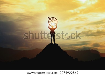 Silhouette of businessman holding a target board on top of mountain. Concept of aim and objective achievement. Royalty-Free Stock Photo #2074922869