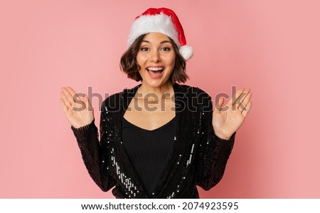 Smiling pretty woman in santa hat  with suprice face celebrating new year party. Posing on pink background.  Stylish outfit.