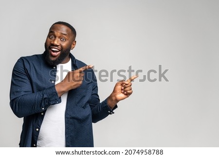 Cheerful overjoyed African-American guy points fingers at empty copy space, isolated on grey background. Multiracial man recommends deal, pays yours attention, presenting new product, advertising