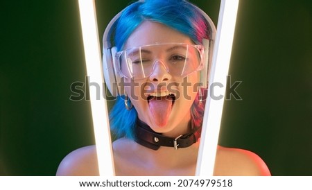 Cyberpunk model. Party girl. Futuristic generation. Funky attractive playful woman sticking tongue out in LED neon light isolated on dark background.