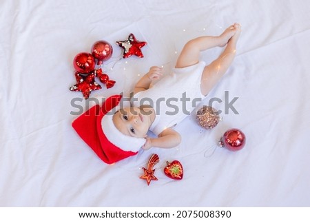 baby in a white bodysuit and a Santa hat is lying on his back on a white sheet surrounded by red Christmas tree toys. winter, new year. space for text. High quality photo