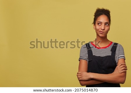 Indoor shot of young african american woman, Wears stripped t shirt and black overalls curving her lips, looks aside at copy space thoughtfully. Isolated over yellow background