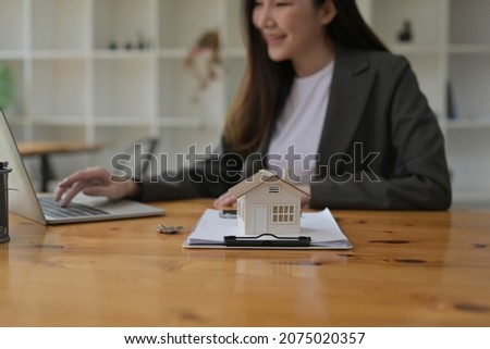 Photo of a house model putting on a clipboard at the wooden working desk over a beautiful woman using a computer laptop as a background.