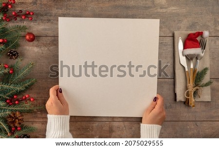 woman hands holds menu in her hands while sitting at Christmas dinner table. Christmas tree decorations border on vintage wooden table with cutlery and mockup christmas menu.