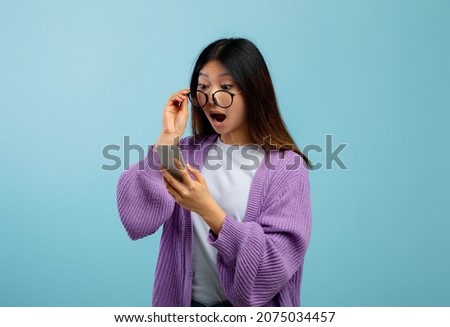 Wow, great news. Shocked asian lady looking at smartphone in excitement, lifting her sunglasses and learning about online seasonal sale, standing over blue background