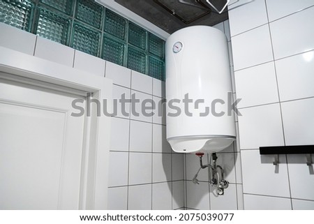 Household budget water heater hanging on the wall in boiler room. Modern gas tanked boiler in bathroom. Common electric storage tank water heater. Energy-efficient home heating system on white tiles Royalty-Free Stock Photo #2075039077