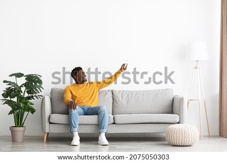 Joyful african american guy in casual sitting on couch at home, holding remote, turning on AC, warming or cooling apartment, full length shot, copy space. Modern technologies at home Royalty-Free Stock Photo #2075050303