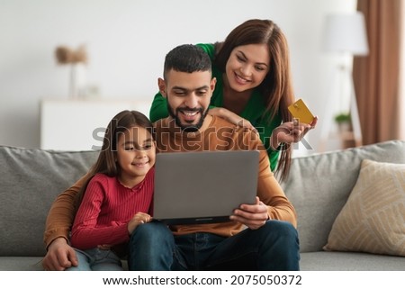Online Shopping Concept. Happy Young Arabic Family With Little Daughter Using Laptop And Debit Credit Card At Home For Buying Goods In Internet Web Store, Three People Sitting On Couch On Living Room Royalty-Free Stock Photo #2075050372