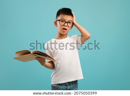 Tired Of Study. Stresses Little Asian Boy Holding Book And Touching Head, Nerdy Korean Male Kid Wearing Eyeglasses Looking At Camera With Frustration, Suffering Learning Problems, Blue Background