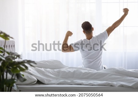 Rear View Of Young Man Stretching In Bed After Waking Up In The Morning, Unrecognizable Male Resting In Light Bedroom After Good Sleep, Looking At Window, Enjoying Start Of New Day, Copy Space Royalty-Free Stock Photo #2075050564