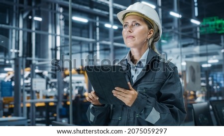 Car Factory Office: Portrait of Female Chief Engineer Wearing Hard Hat Monitoring Production Conveyor with Tablet Computer. Automated Robot Arm Assembly Line Manufacturing High-Tech Electric Vehicles Royalty-Free Stock Photo #2075059297