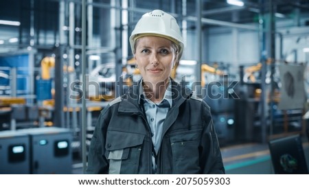 Car Factory Office: Portrait of Female Chief Engineer Wearing Hard Hat Looking at Camera, Smiling. Professional Technician. Automated Robot Arm Assembly Line Manufacturing High-Tech Electric Vehicles Royalty-Free Stock Photo #2075059303