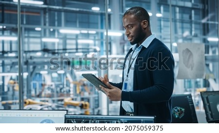 Car Factory Office: Portrait of Successful Black Male Chief Engineer Using Tablet Computer in Automated Robot Arm Assembly Line Manufacturing High-Tech Electric Vehicles. Side View Shot Royalty-Free Stock Photo #2075059375