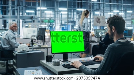 Car Factory Office: Male Automotive Engineer Sitting at His Desk Working on Green Screen Chroma Key Computer. Automated Robot Arm Assembly Line Manufacturing. Medium Shot