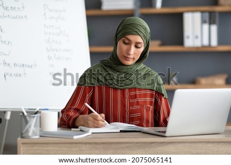 Online Education. Portrait of muslim woman in headscarf sitting at table writing in notebook with pen, using pc computer having video training during quarantine due to covid-19 coronavirus pandemic