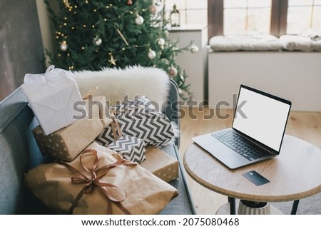 Laptop on table in interior with Christmas tree and wrapped presents. Holiday online shopping.